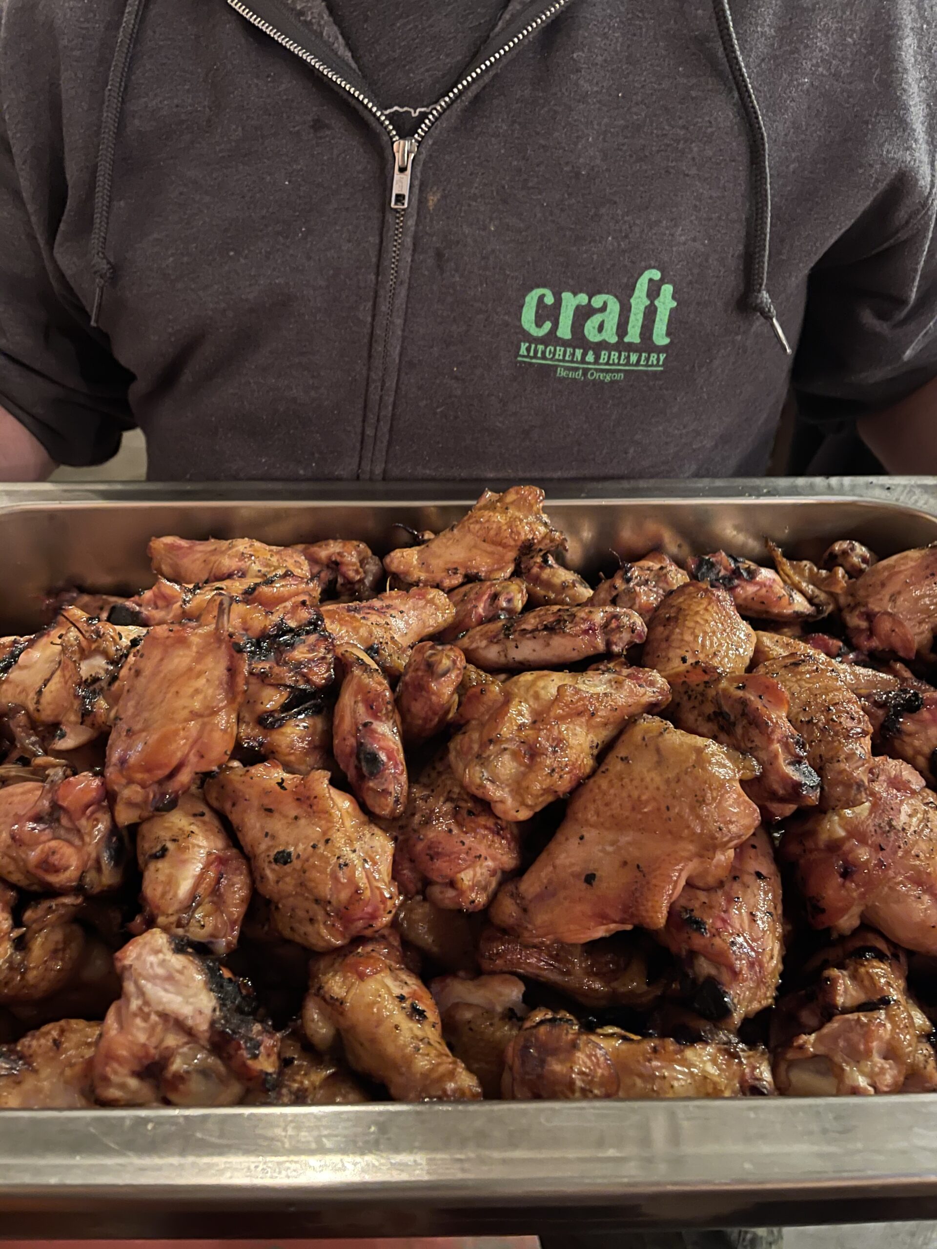A man in a Craft Kitchen & Brewery Hoodie holding a pan of barbeque chicken ready to eat